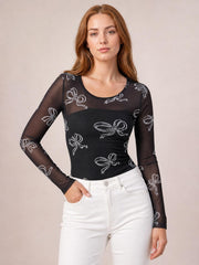 Model poses with one hand in her pocket wearing a black sheer bow-print round-neck top with fitted long sleeves and a slim silhouette.