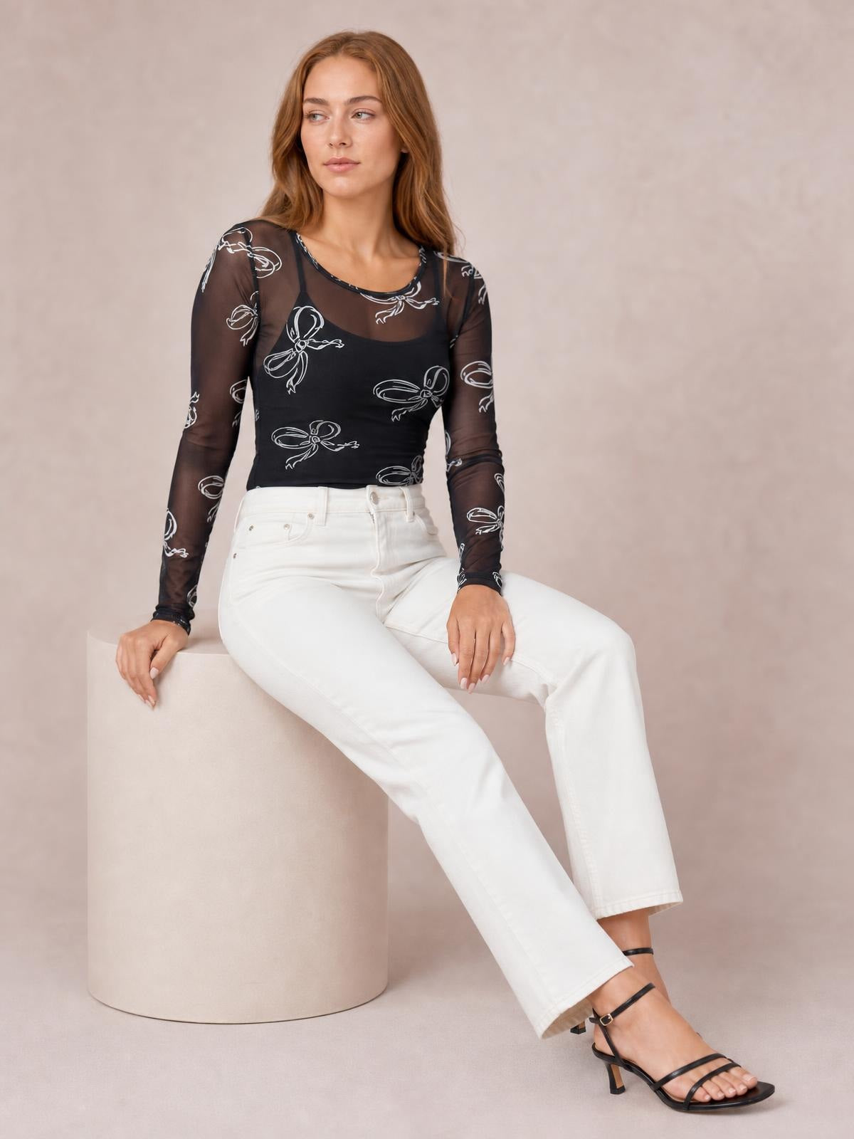 A seated model wears a black sheer round-neck long-sleeve top with a fitted silhouette and white bow print throughout.