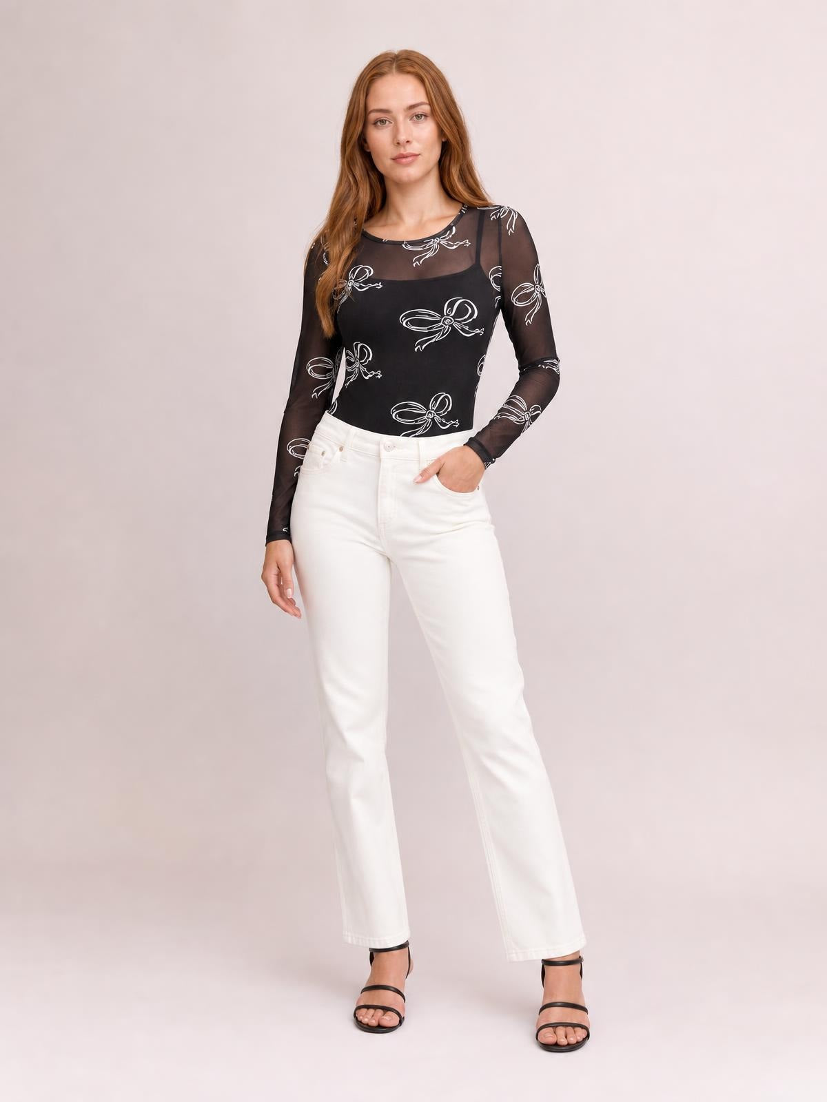 Model stands in a fitted black sheer round-neck long-sleeve top with white bow prints, paired with white straight-leg pants and one hand in a pocket.