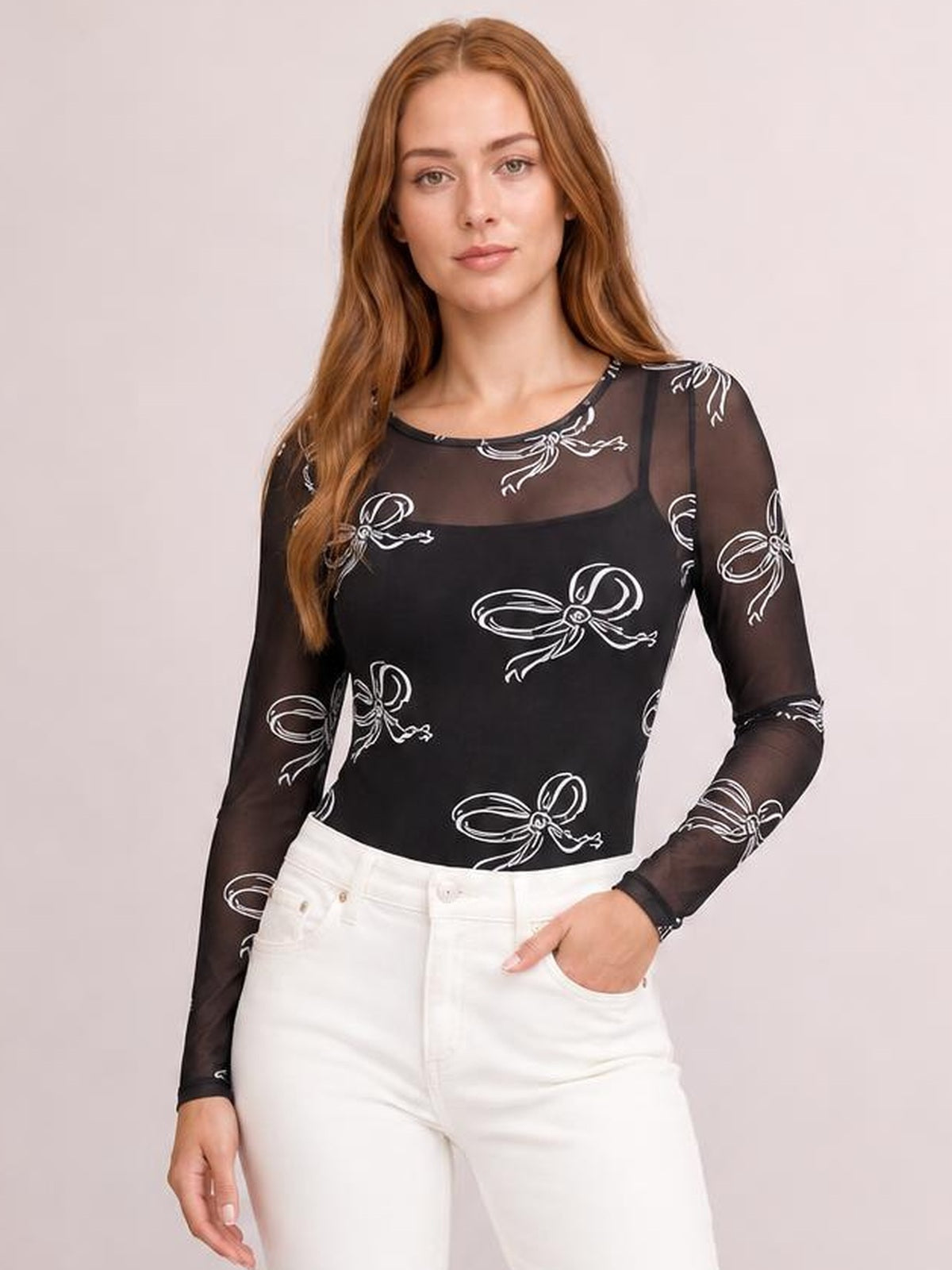 Model poses with one hand in her pocket wearing a fitted black sheer round-neck long-sleeve top with white bow prints and slightly stretchy construction.