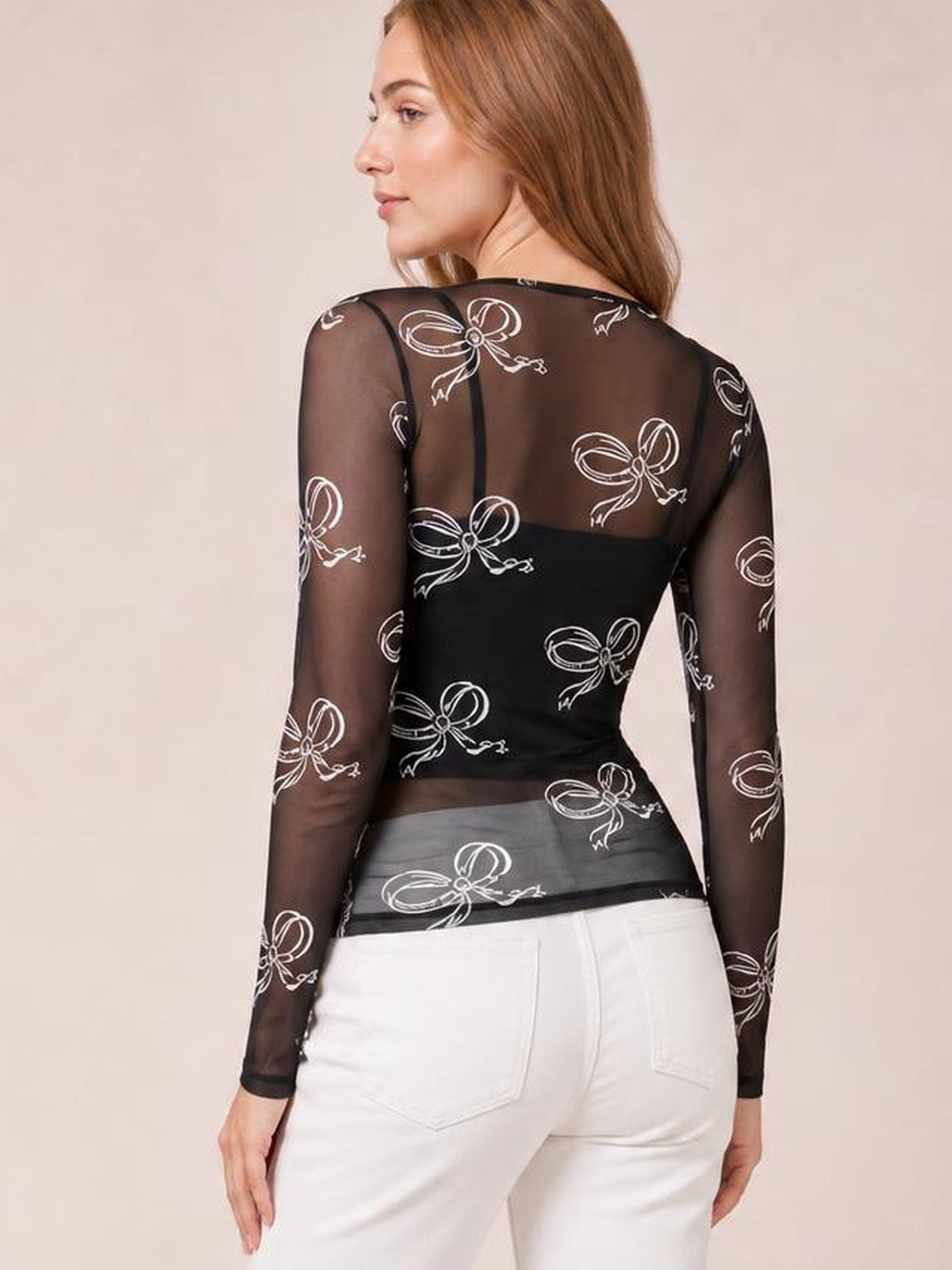 A woman poses with her back turned in a black sheer long-sleeve top featuring a round neckline, slim silhouette, and white bow print.