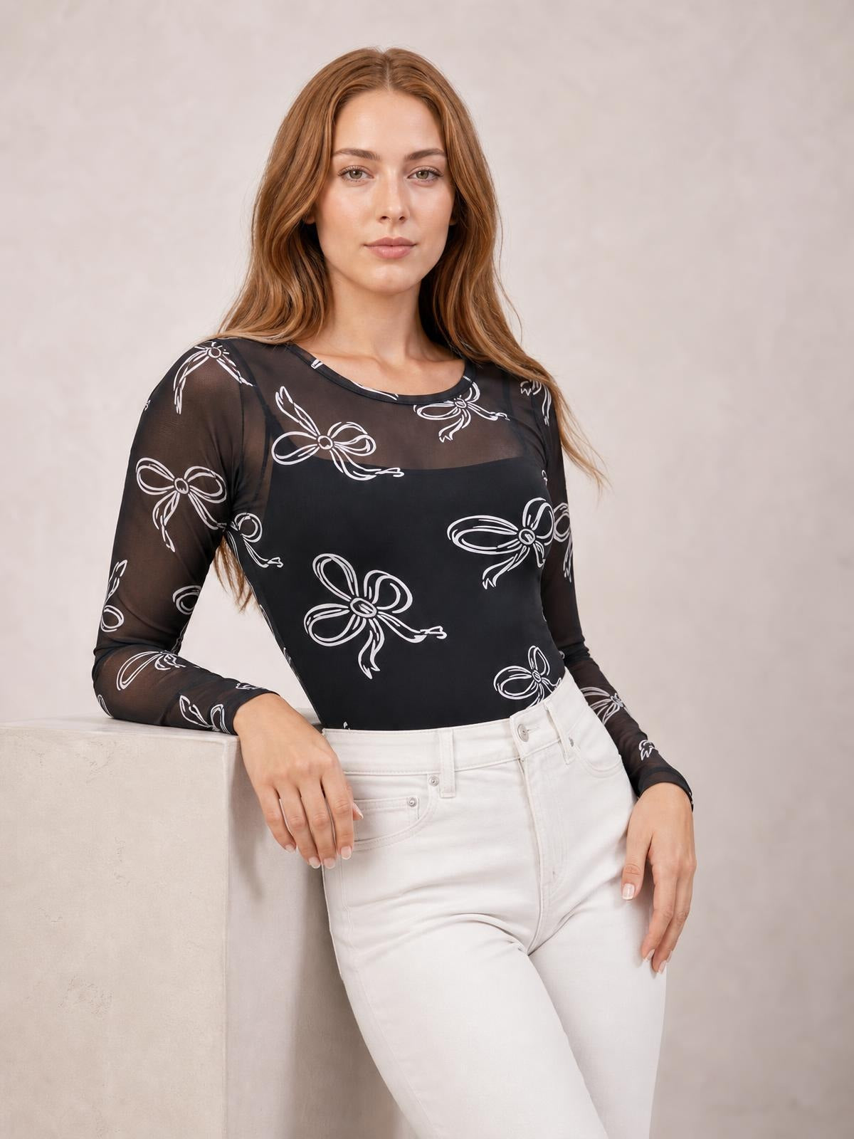A model poses in a fitted black sheer round-neck long-sleeve top with white bow print, resting one arm on a ledge and showing its sleek silhouette.