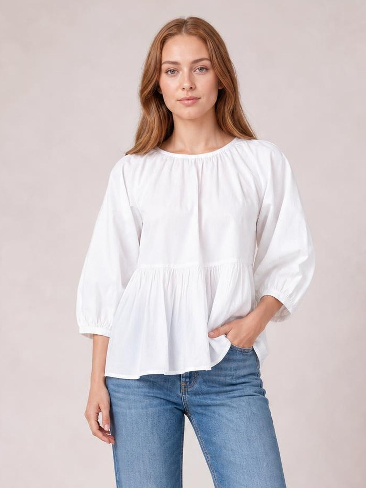 A model poses in a white round-neck blouse with three-quarter sleeves, gathered waist, and a softly flared peplum silhouette.