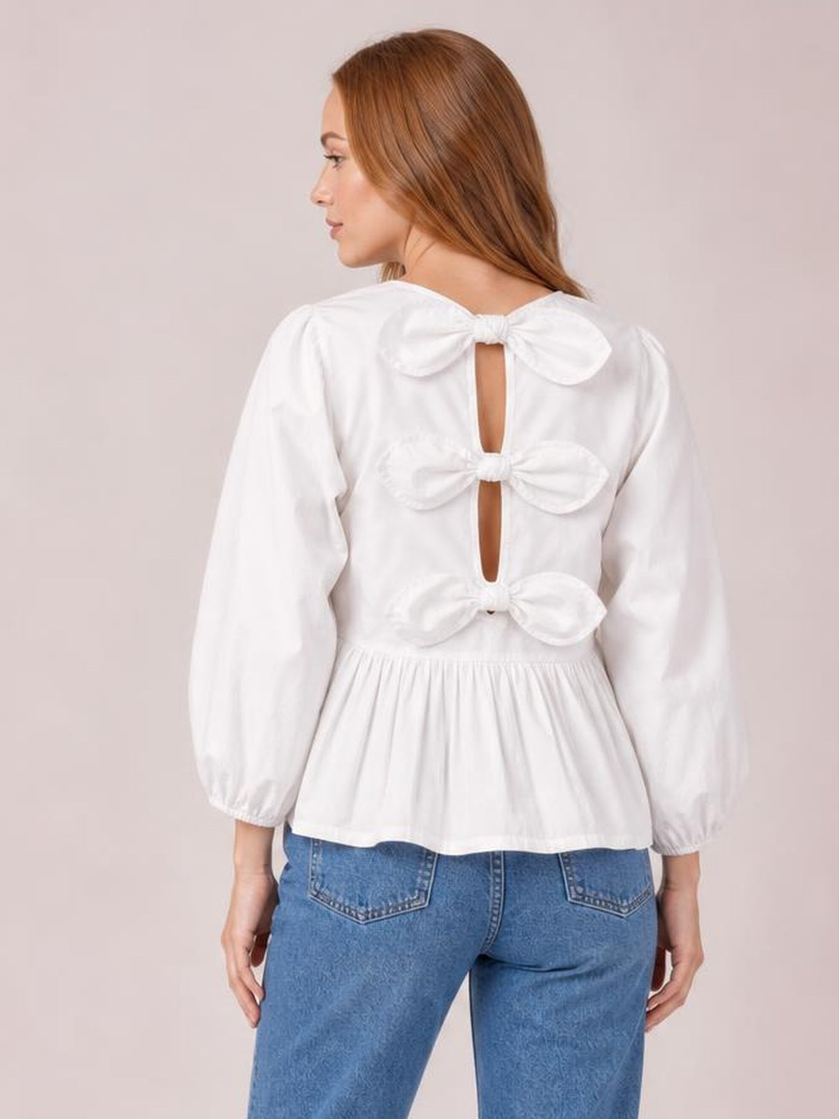 Back view of a model wearing a white bow blouse with a round neckline, three-quarter sleeves, three back bows with cutouts, and a gathered peplum silhouette.