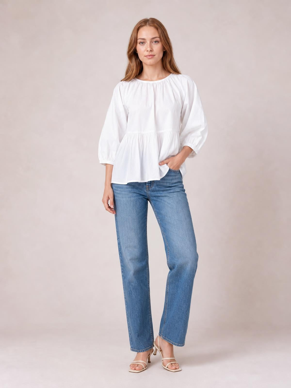 Model wears a white round-neck blouse with gathered detailing, three-quarter sleeves, and a softly flared peplum silhouette while posing with one hand in her jeans pocket.