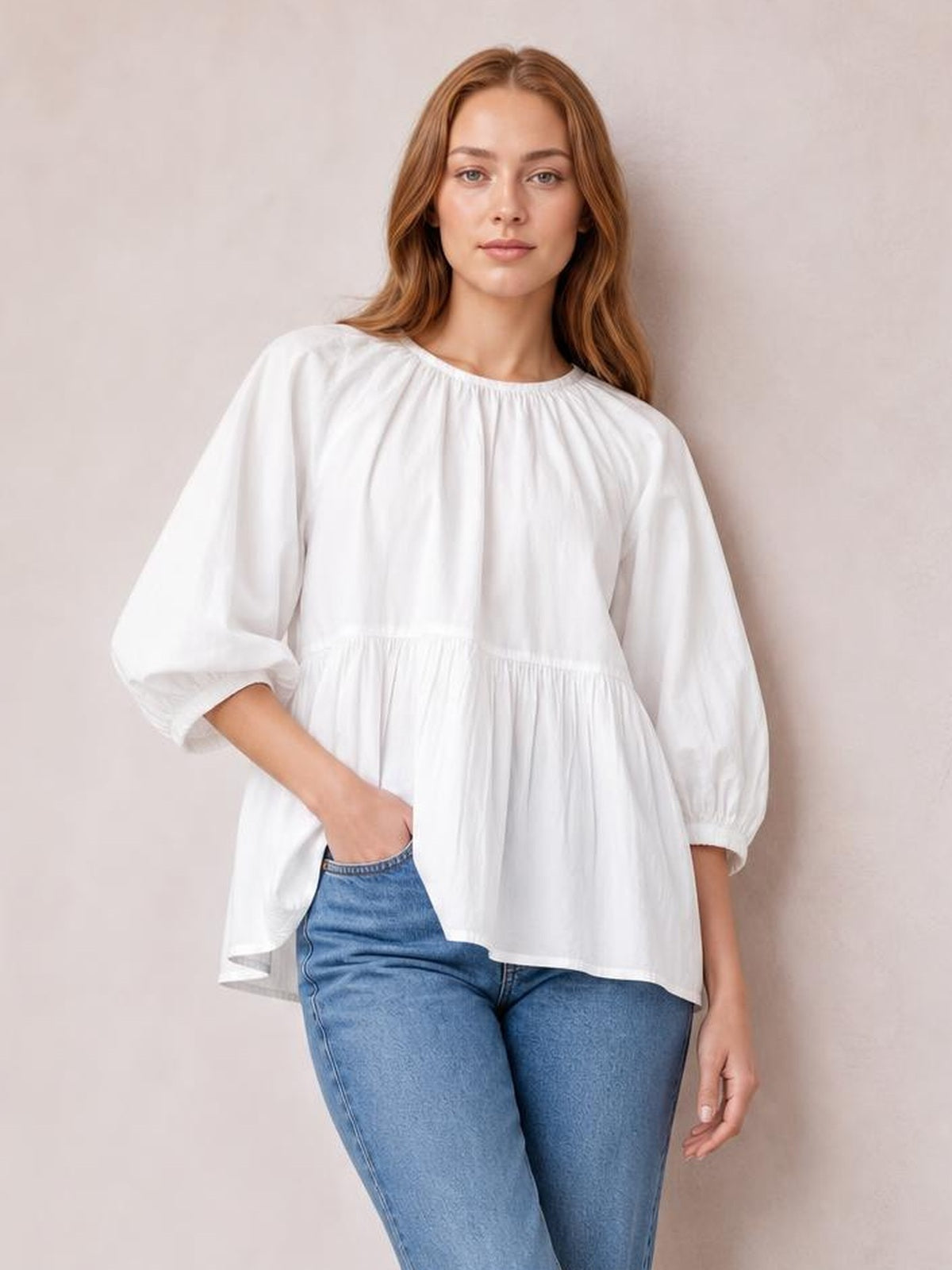 Model wears a white round-neck blouse with three-quarter sleeves, gathered construction, a relaxed flared silhouette, and one hand resting in her jeans pocket.
