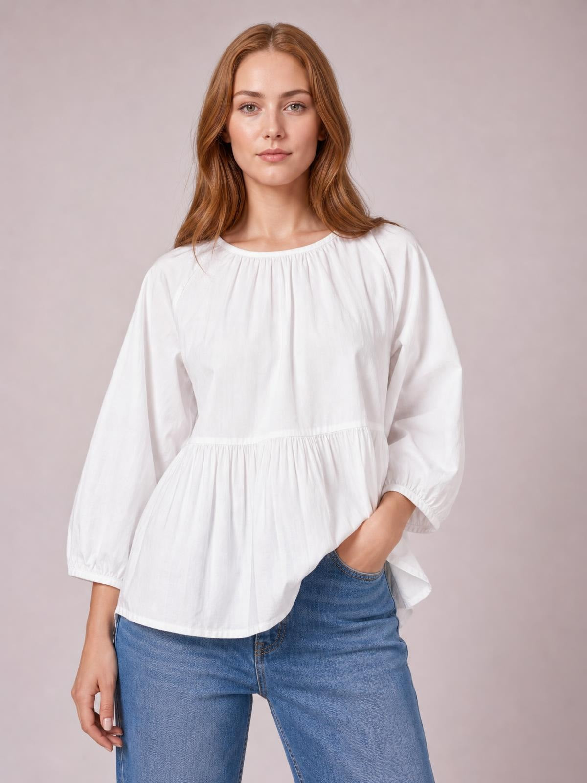 A model wears a white round-neck blouse with three-quarter sleeves, gathered construction, and a relaxed peplum silhouette, posing with one hand in her jeans pocket.