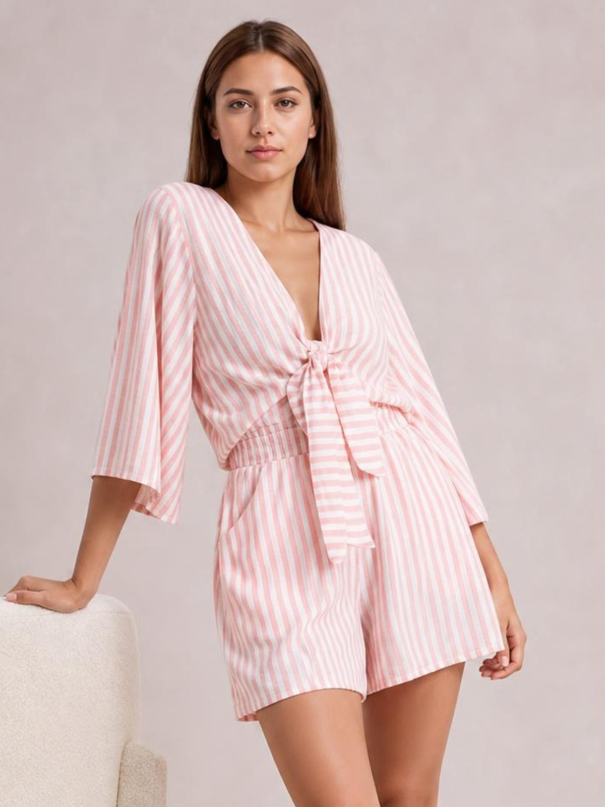 Model wearing a pink-and-white striped romper with kimono sleeves, a tied waist bow, V-neckline, elastic waist, and relaxed shorts silhouette.