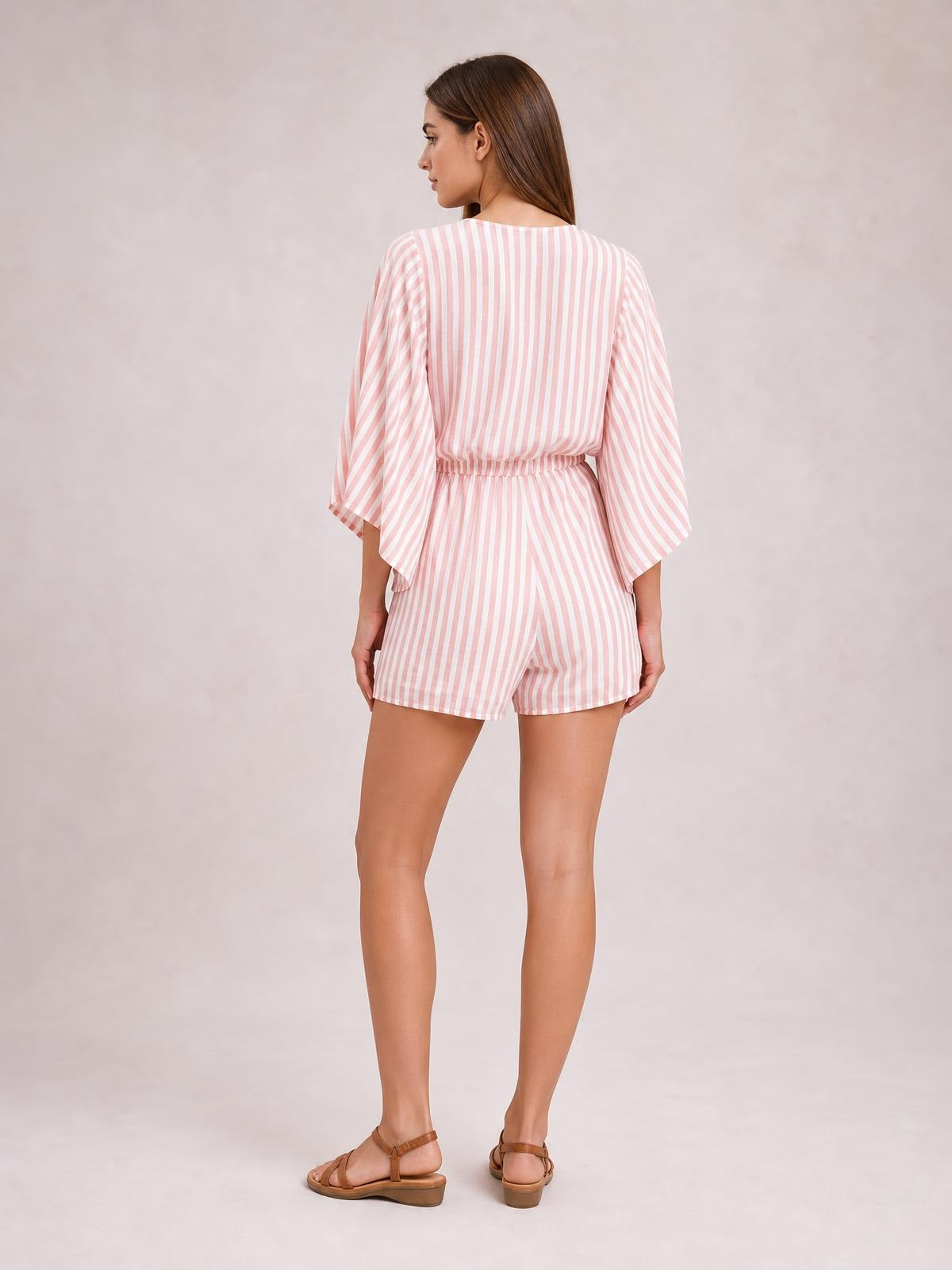 Model shown from the back wearing a pink-and-white striped romper with loose kimono sleeves, an elastic waist, and relaxed shorts silhouette.