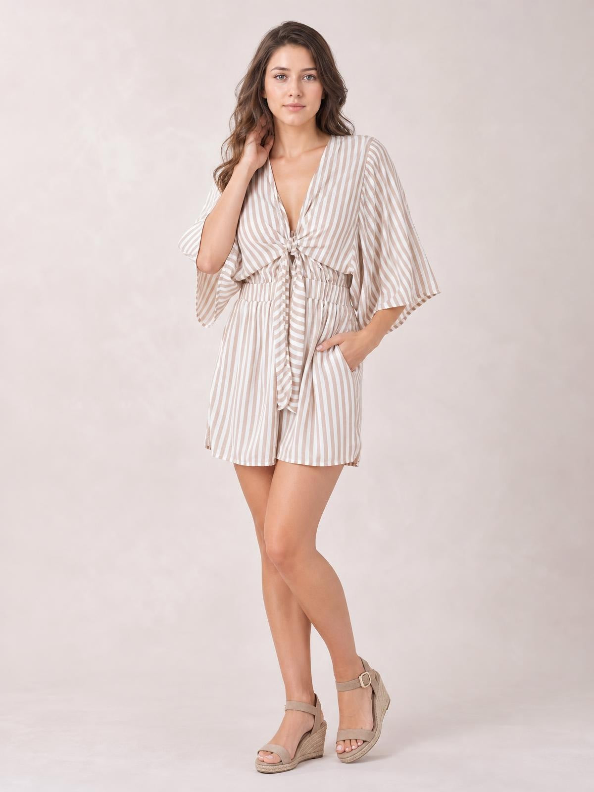 Model poses in a beige-and-white striped romper with flowing kimono sleeves, a tied front bow, elastic waist, and relaxed shorts silhouette.