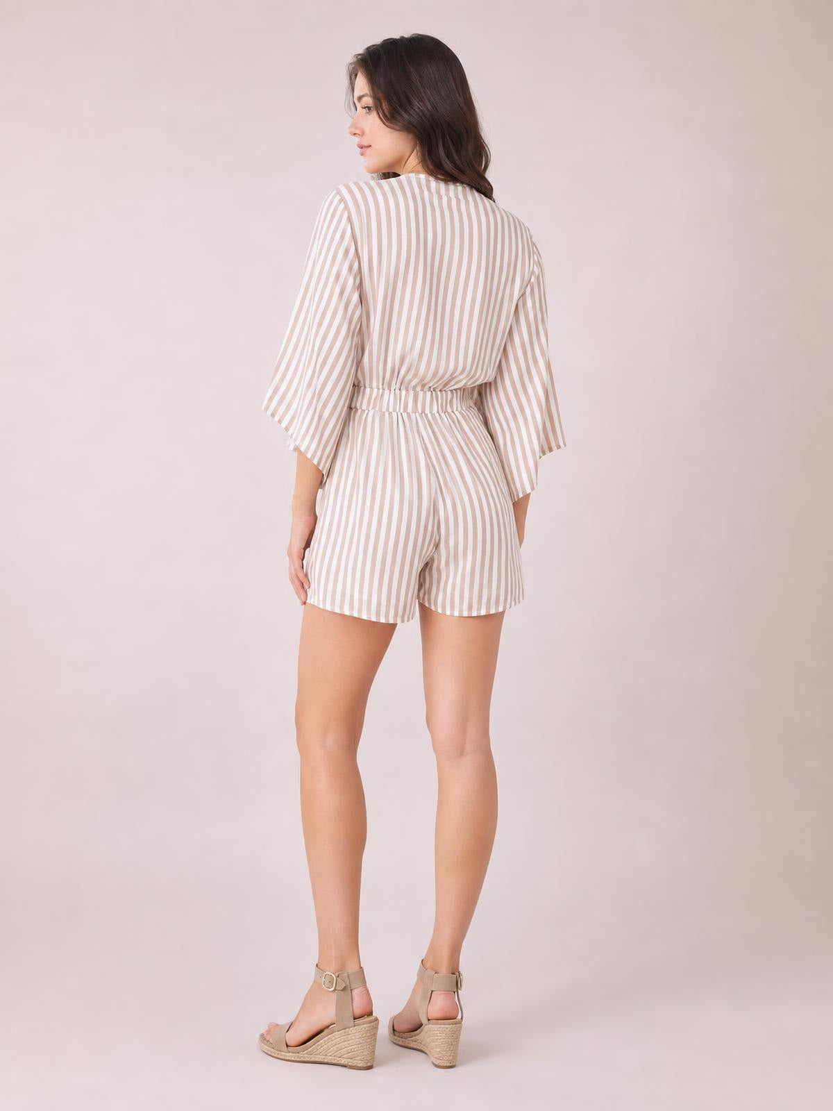 Model shown from the back in a beige-and-white striped romper with kimono sleeves, tied waist bow, elastic waistband, and relaxed shorts silhouette.