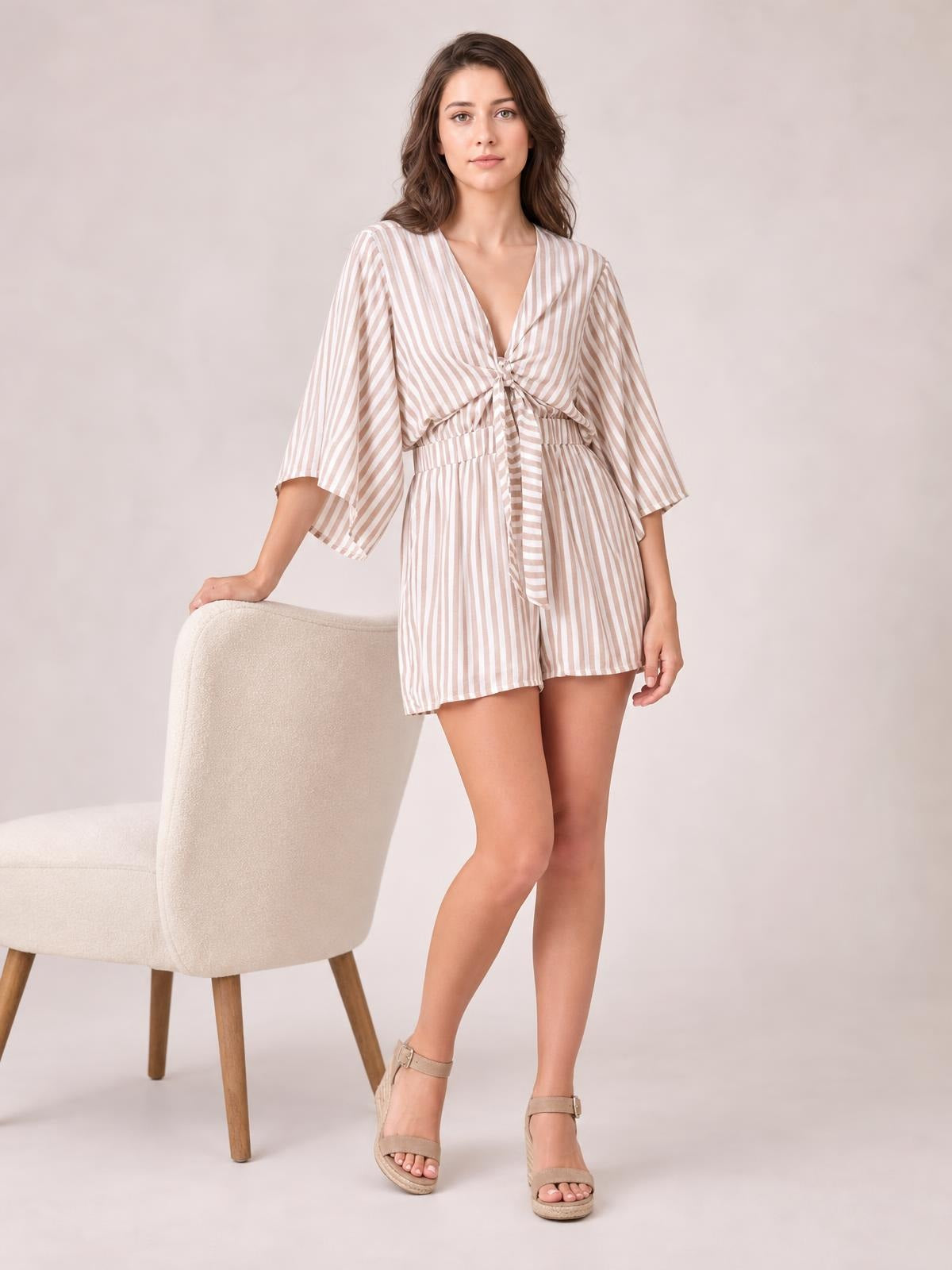 Model wears a beige-and-white striped romper with flowing kimono sleeves, a tied front bow, elastic waist, and relaxed shorts, posing with one hand on a chair.