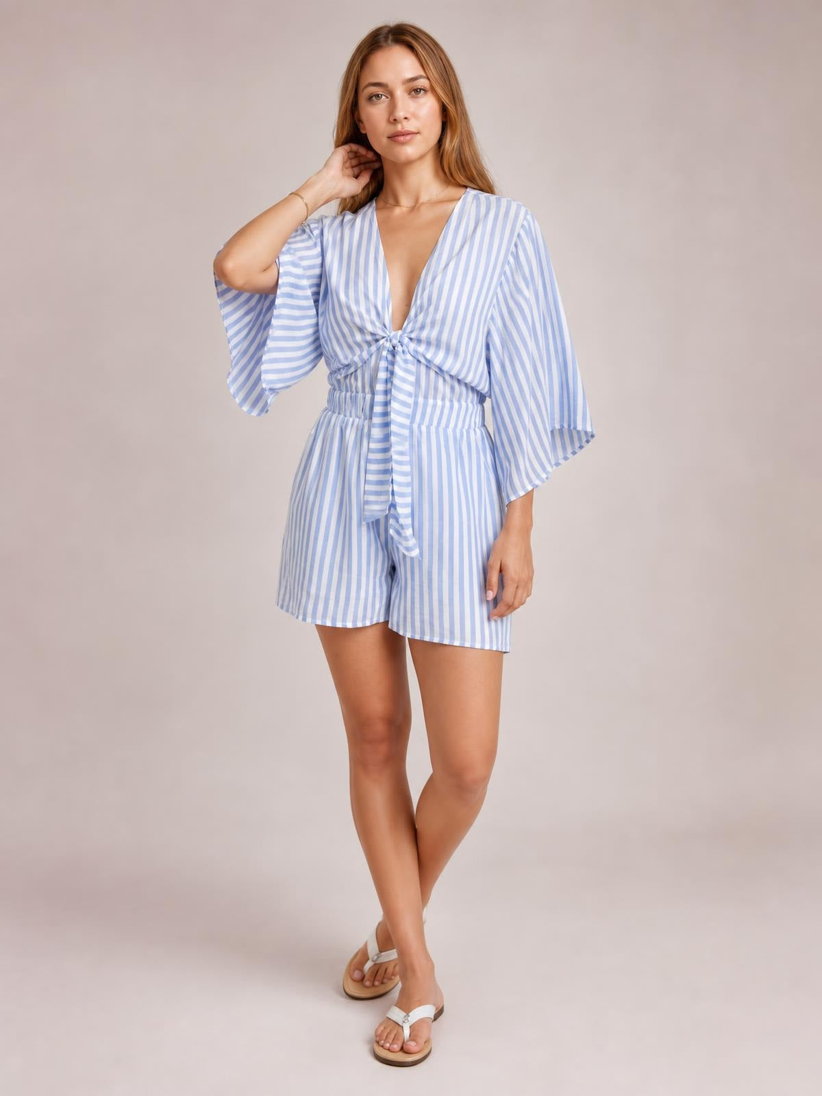 Model poses in a light blue and white striped romper with kimono sleeves, a tied front bow, elastic waist, and relaxed shorts silhouette.