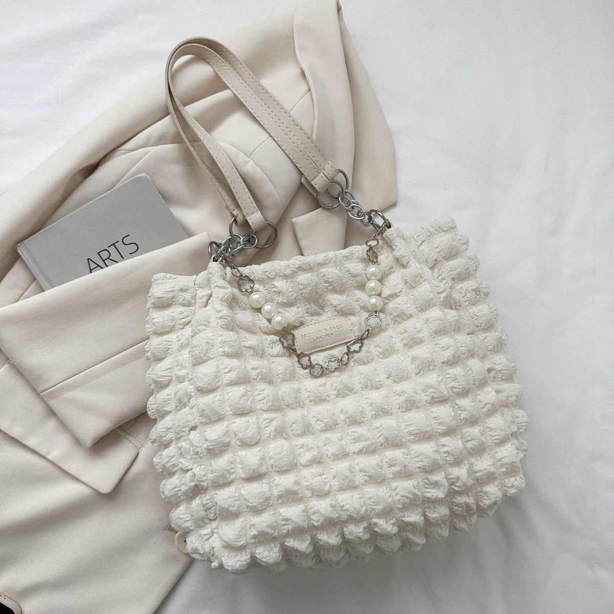 A white medium Bubble Textured Tote Bag features a raised dimensional surface, slender straps, linked chain accents, a pearl-style strand, and a front plaque.