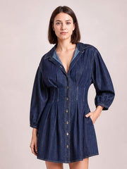 Model poses in a dark blue denim dress with a button-down front, collared neckline, 3/4 sleeves, seam detailing, fitted waist, and flared skirt.