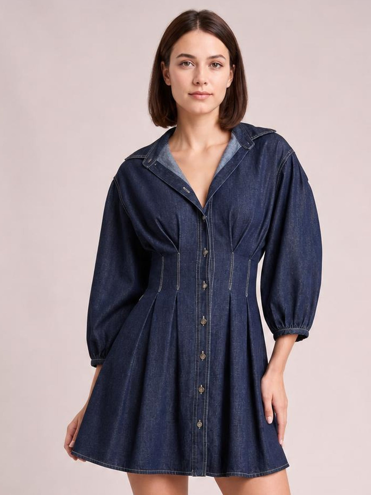 Model poses in a dark blue button-down denim dress with seam detailing, a collared neckline, gathered 3/4 sleeves, fitted waist, and flared silhouette.