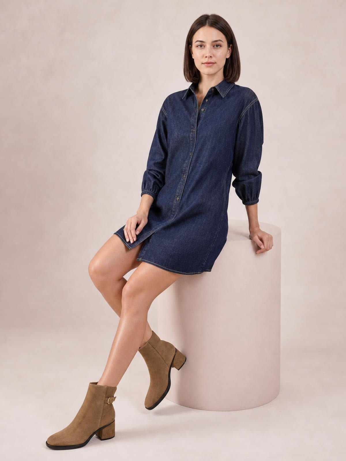 A seated model wears a dark blue button-down denim dress with a collared neckline, 3/4 sleeves, visible seam detail, and a relaxed knee-length silhouette.