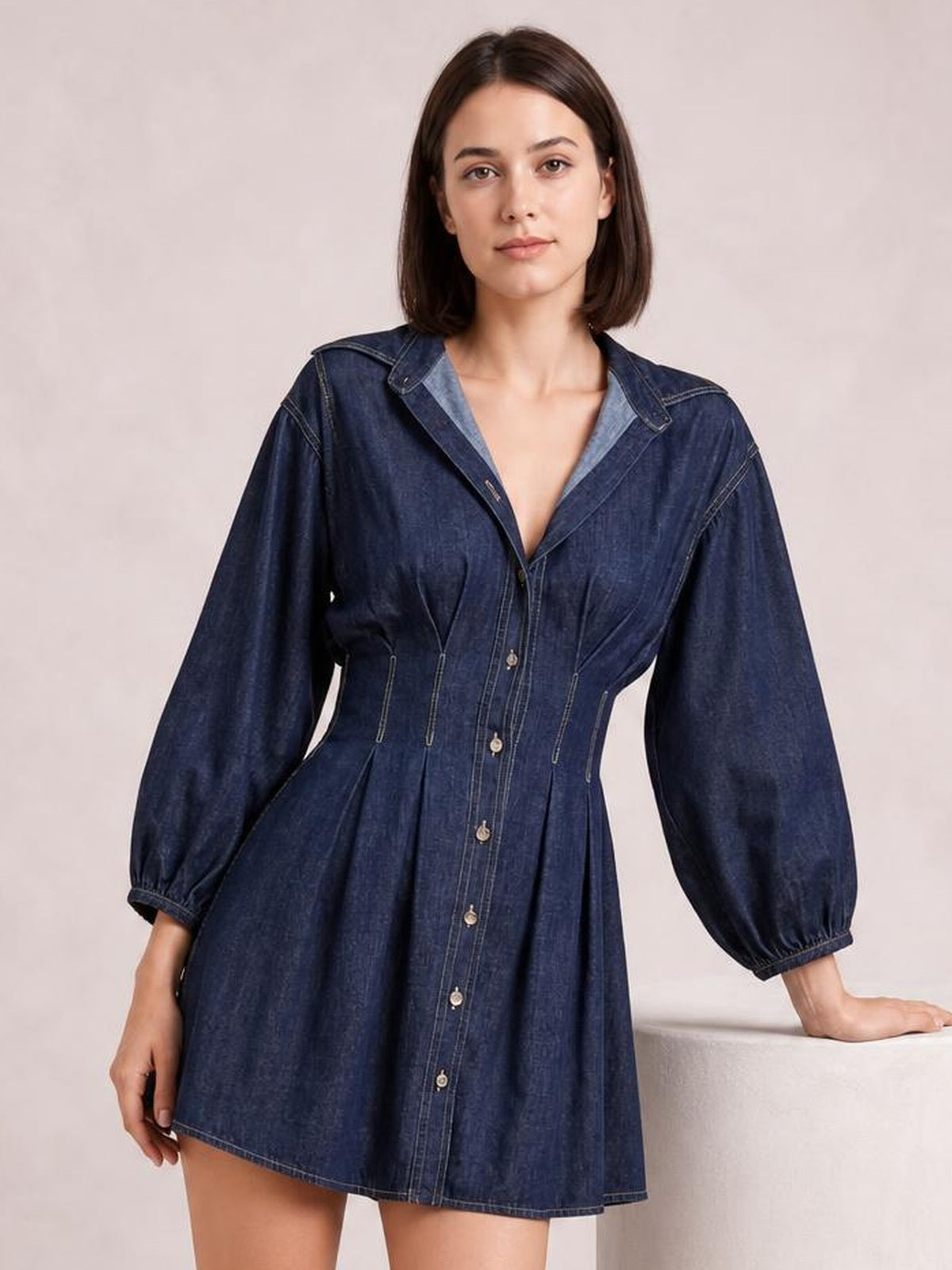 A model poses in a dark blue button-down denim dress with 3/4 sleeves, contrast seam details, a fitted waist, and a pleated flared skirt.