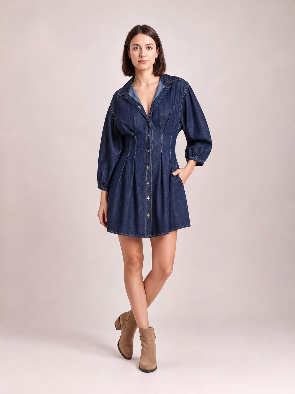 A model poses in a dark blue button-down denim dress with a collared neckline, 3/4 sleeves, seam detailing, fitted waist, and flared short skirt.
