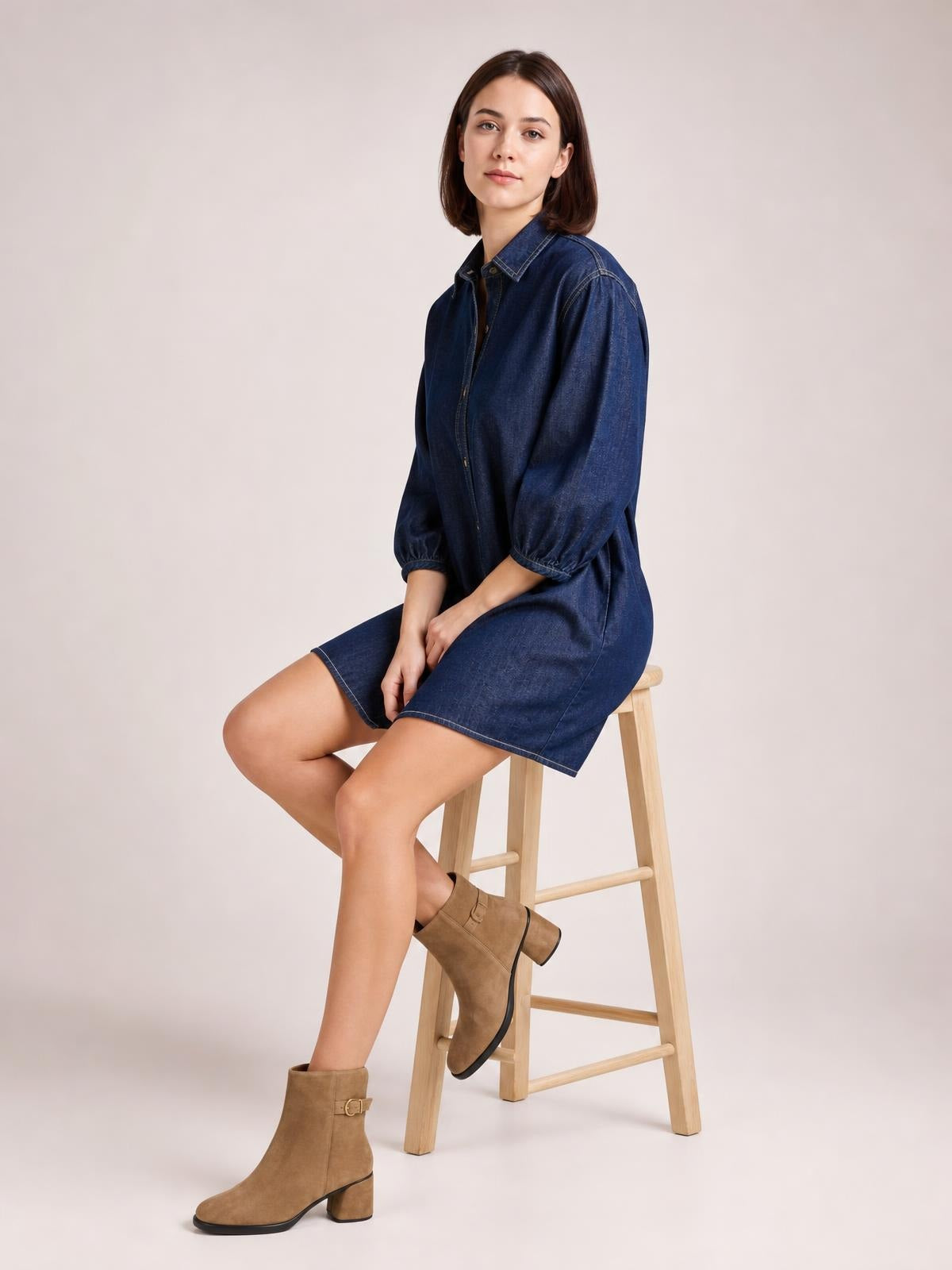 Model seated in a relaxed pose wears a dark blue button-down denim dress with a collared neckline, visible seam details, 3/4 sleeves, and a straight mini silhouette.
