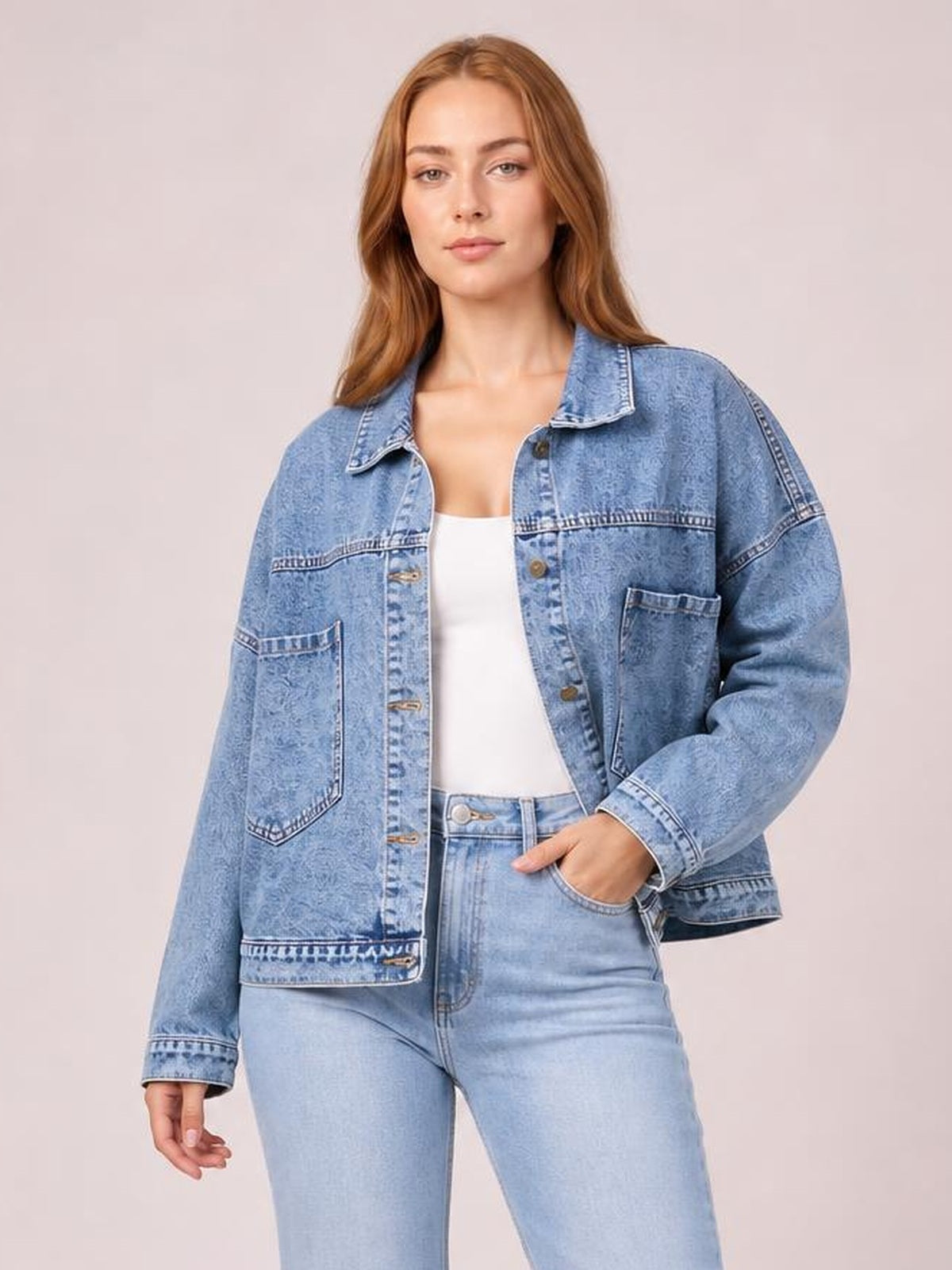 A model poses in a light blue button-down denim jacket with drop shoulders, long sleeves, a relaxed silhouette, and two front patch pockets.