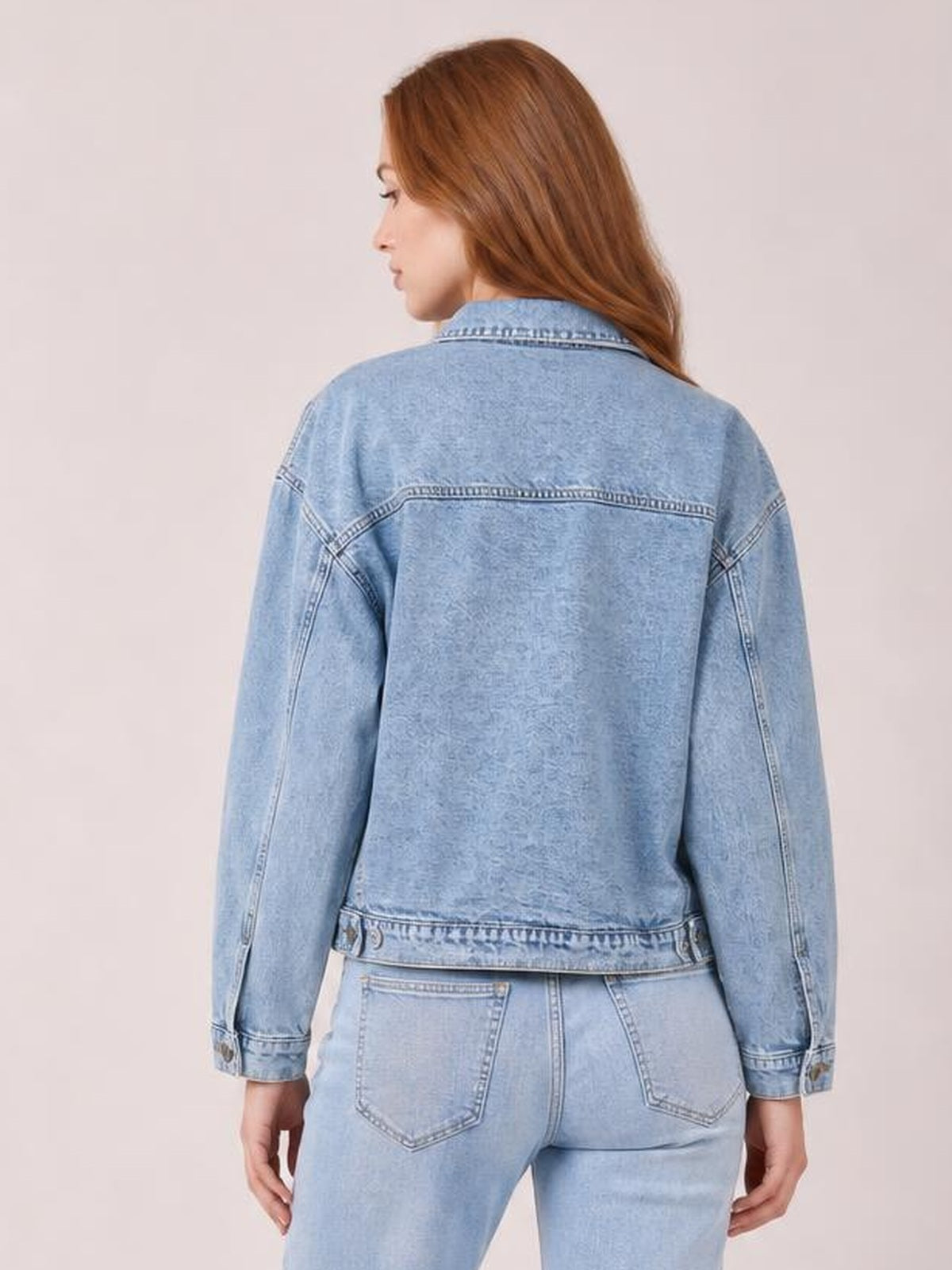 A model stands facing away in a light blue denim jacket with drop shoulders, long sleeves, a collared neckline, seam detailing, and a relaxed straight silhouette.