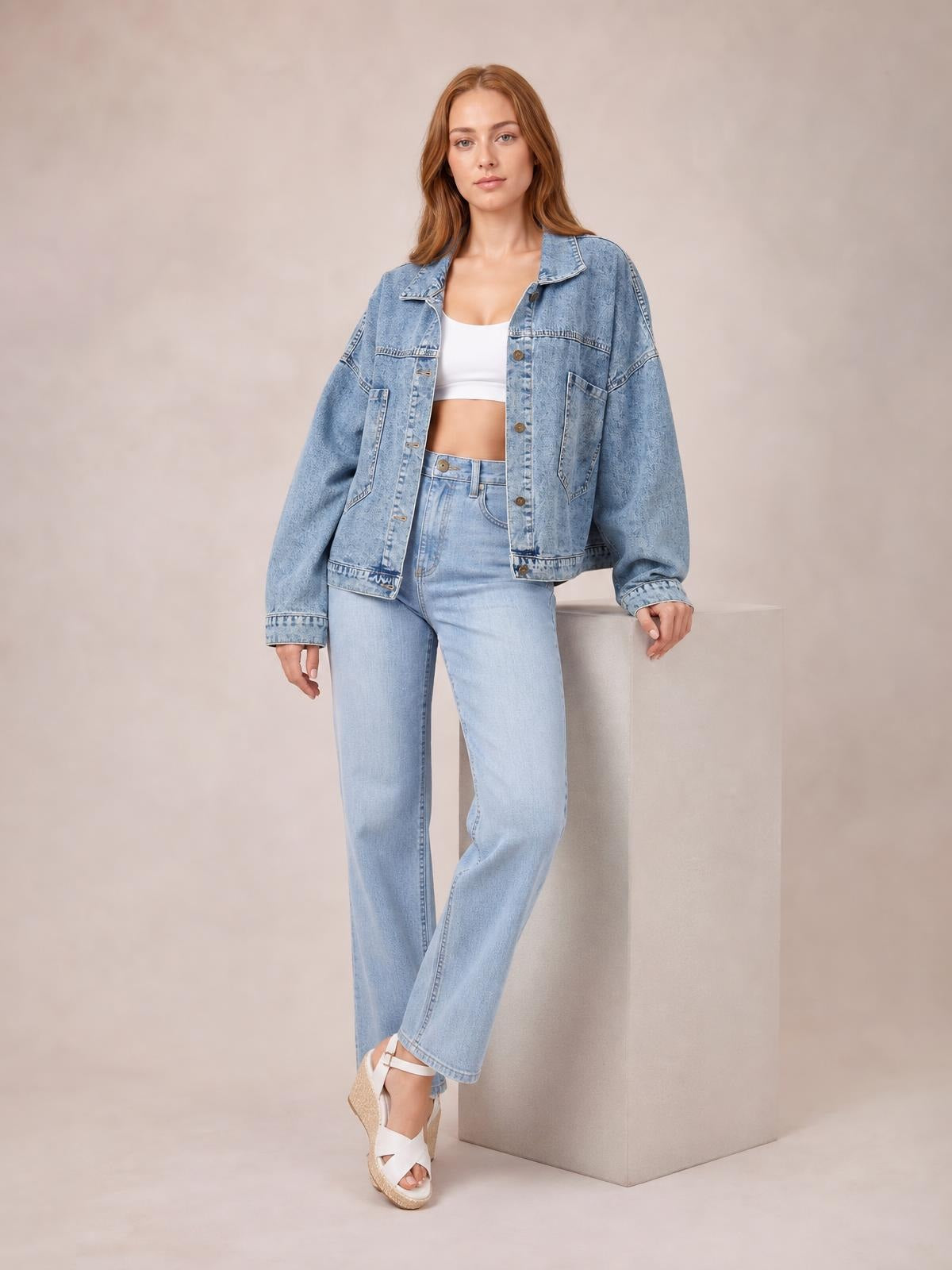 A model wears a light blue button-down denim jacket with a relaxed drop-shoulder silhouette, long sleeves, collar, and front pockets, posing with one leg crossed.
