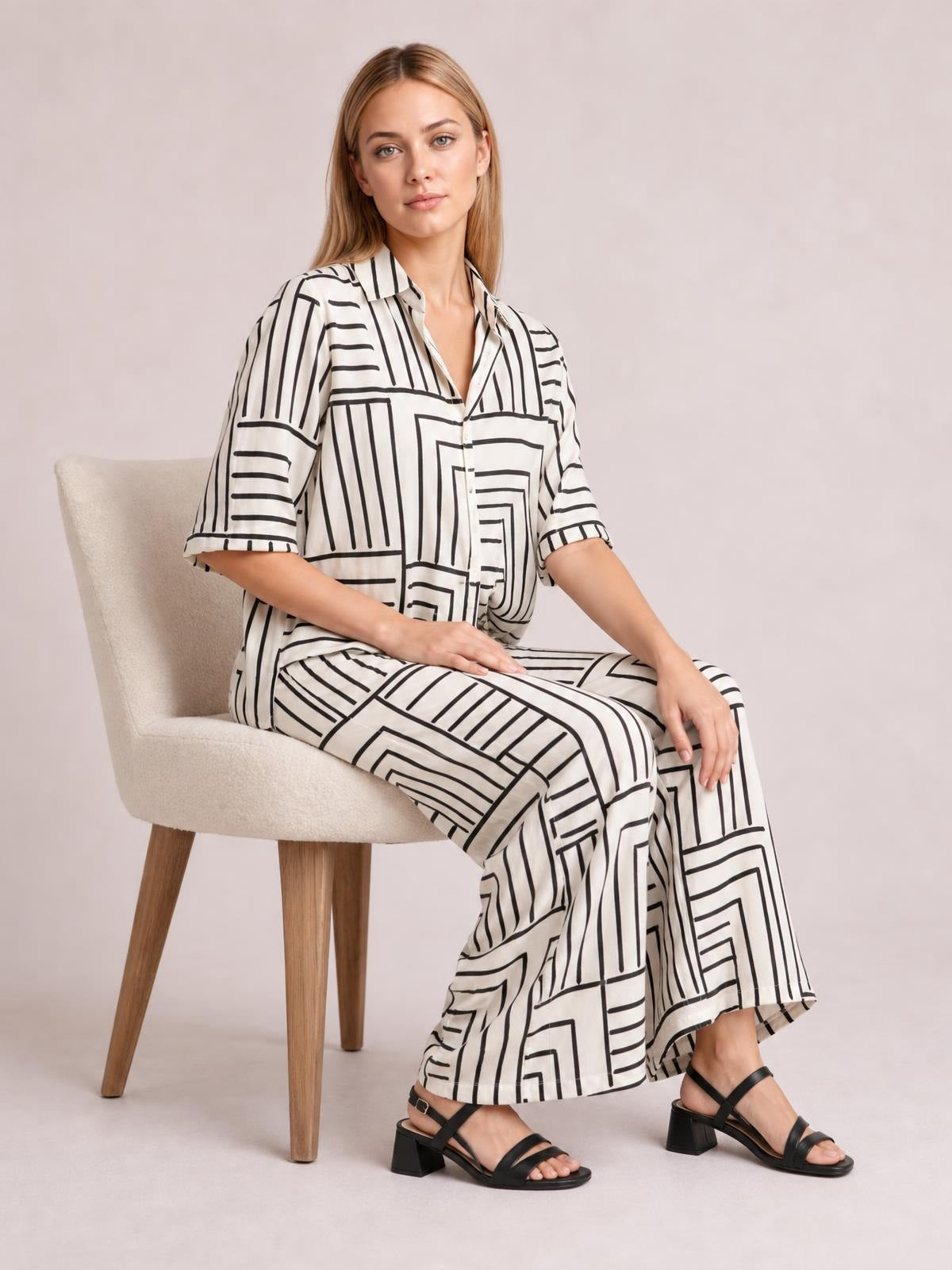 A seated model wears a white button-down half-sleeve top with black geometric stripes and a chest pocket, paired with matching wide-leg pants.