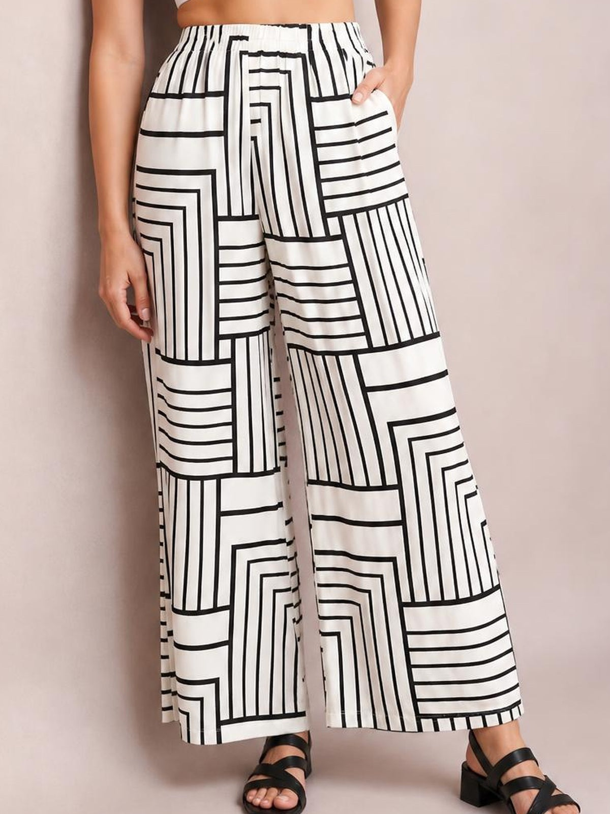 A model wears white wide-leg pants with bold black geometric line patterns, a gathered waistband, and side pockets, standing with one hand tucked in a pocket.