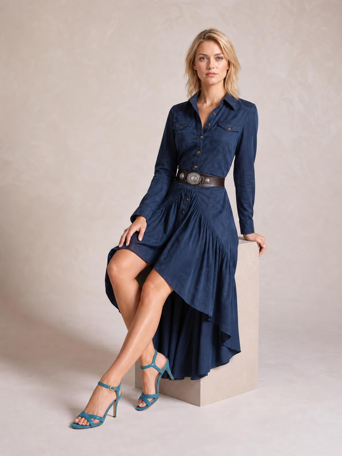 A model sits in a navy button-down shirt dress with a collared long-sleeve bodice, chest pockets, and a flowing ruffled high-low hem.