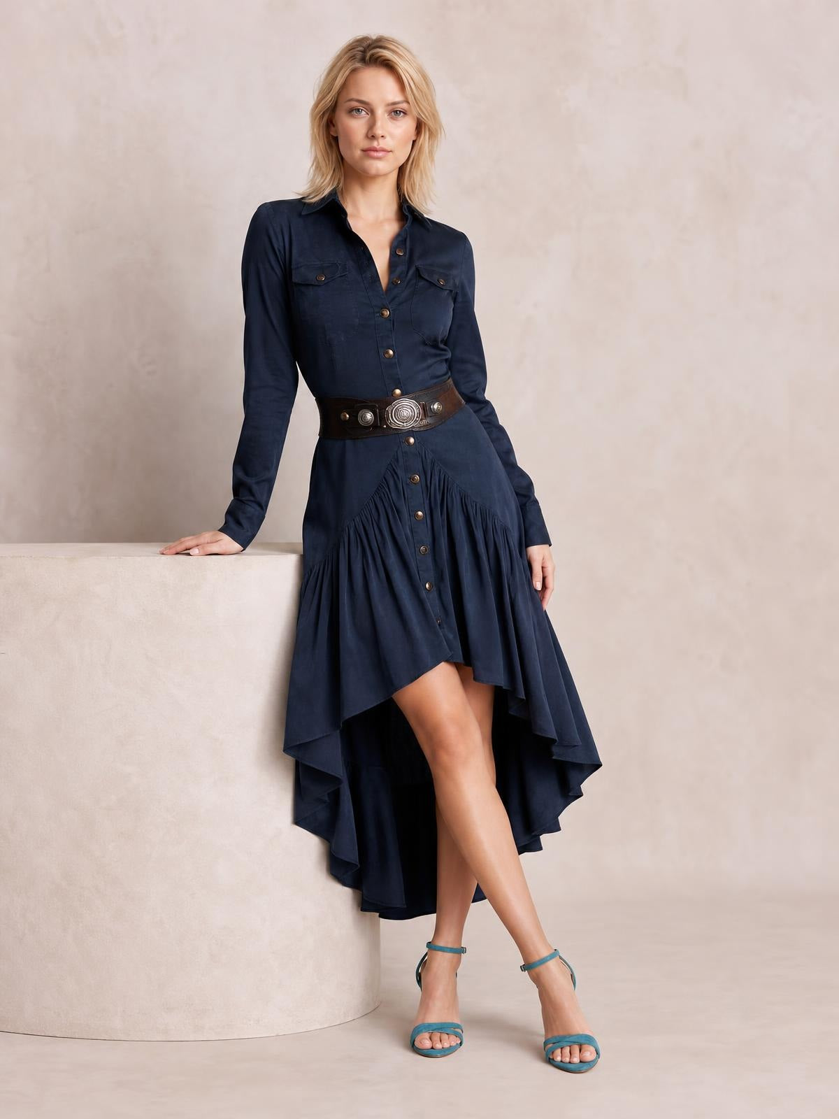 A model leans in a navy button-down shirt dress with long sleeves, flap pockets, a belted waist, and a ruffled high-low hem.