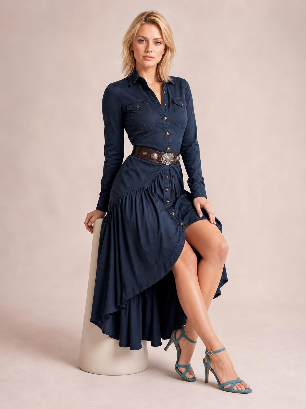 A model sits in a navy button-down shirt dress with a collared neckline, long sleeves, chest pockets, gathered ruffles, and a flowing high-low hem.