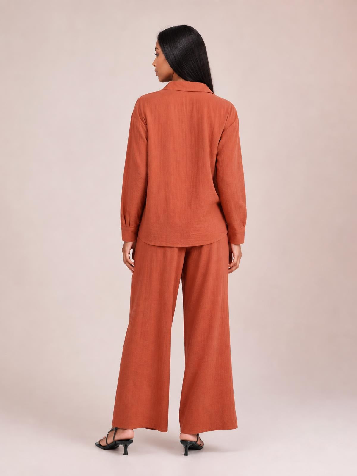 Model shown from behind in a rust-colored long-sleeve button-down top with cuffed sleeves and matching wide-leg pants, standing in black heels.
