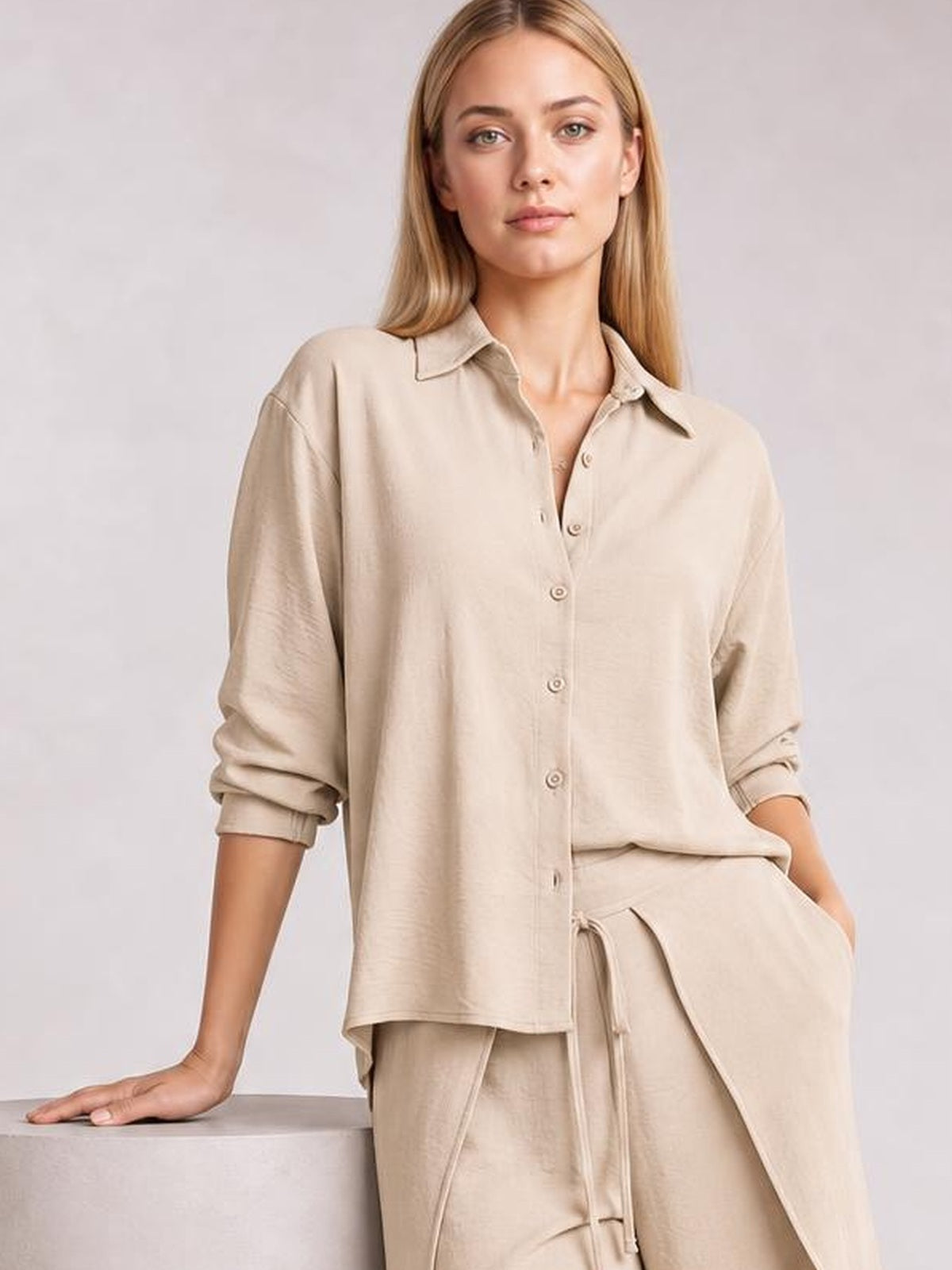 A model leans against a surface wearing a beige button-down long-sleeve top with rolled cuffs and coordinating wide-leg pants with a drawstring waist.