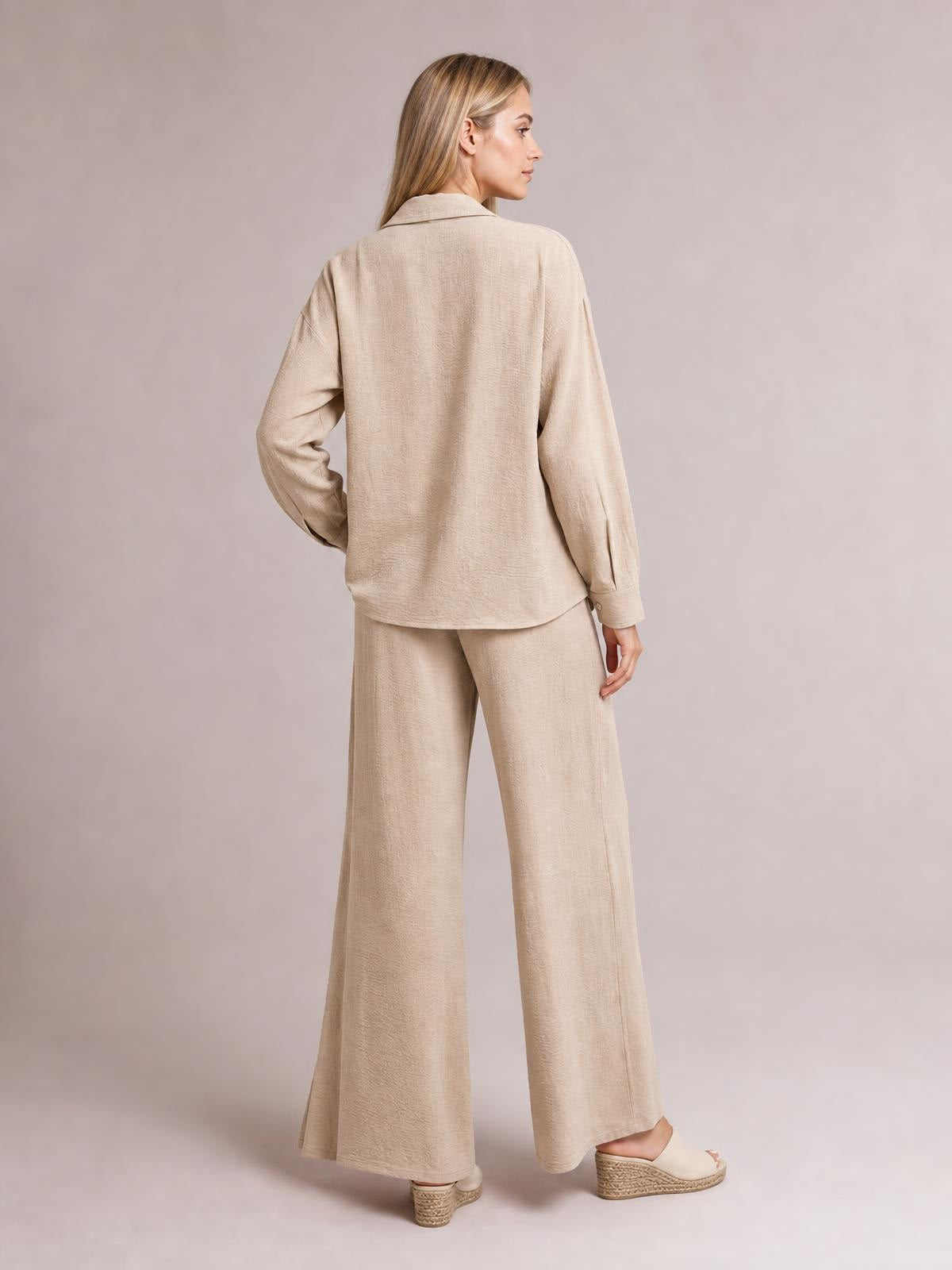 Model in a beige button-down long-sleeve top and matching wide-leg pants set, shown from behind in a relaxed pose with a fluid, tailored silhouette.