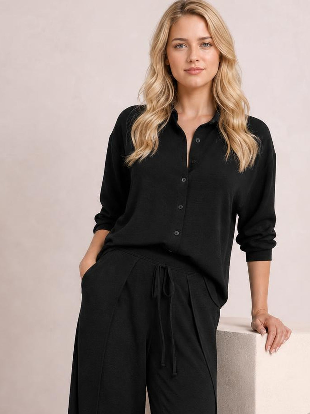 A model leans against a ledge wearing a black button-down long-sleeve top and matching wide-leg pants with a visible drawstring waist.