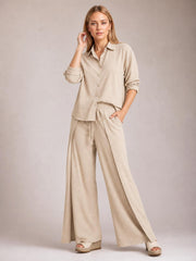 A model poses in a beige button-down long-sleeve top and wide-leg pants set, featuring a relaxed silhouette, drawstring waist, and side slit accents.