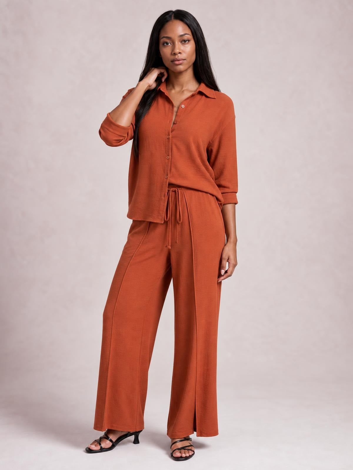A model poses in a rust-colored button-down long-sleeve top and wide-leg pants set with a relaxed silhouette, drawstring waist, and slit hems.
