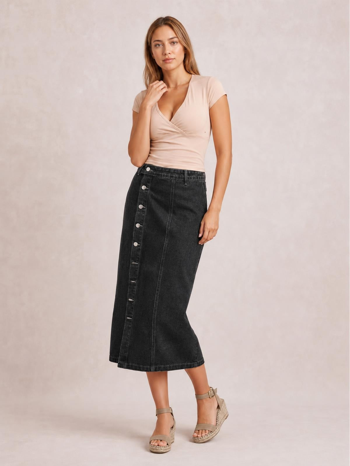 A model poses with one hand near her neckline, wearing a dark denim midi skirt with a straight silhouette, waistband, seams, and button-down front.