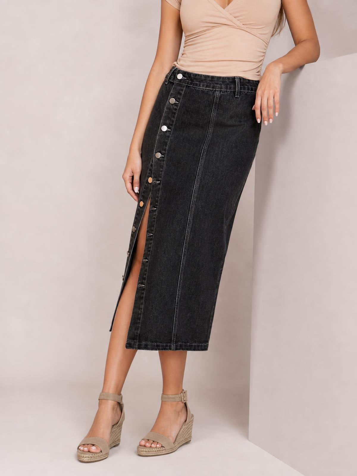 A model leans against a wall wearing a dark denim midi skirt with a straight silhouette, button-down front, front slit, waistband, and seam details.