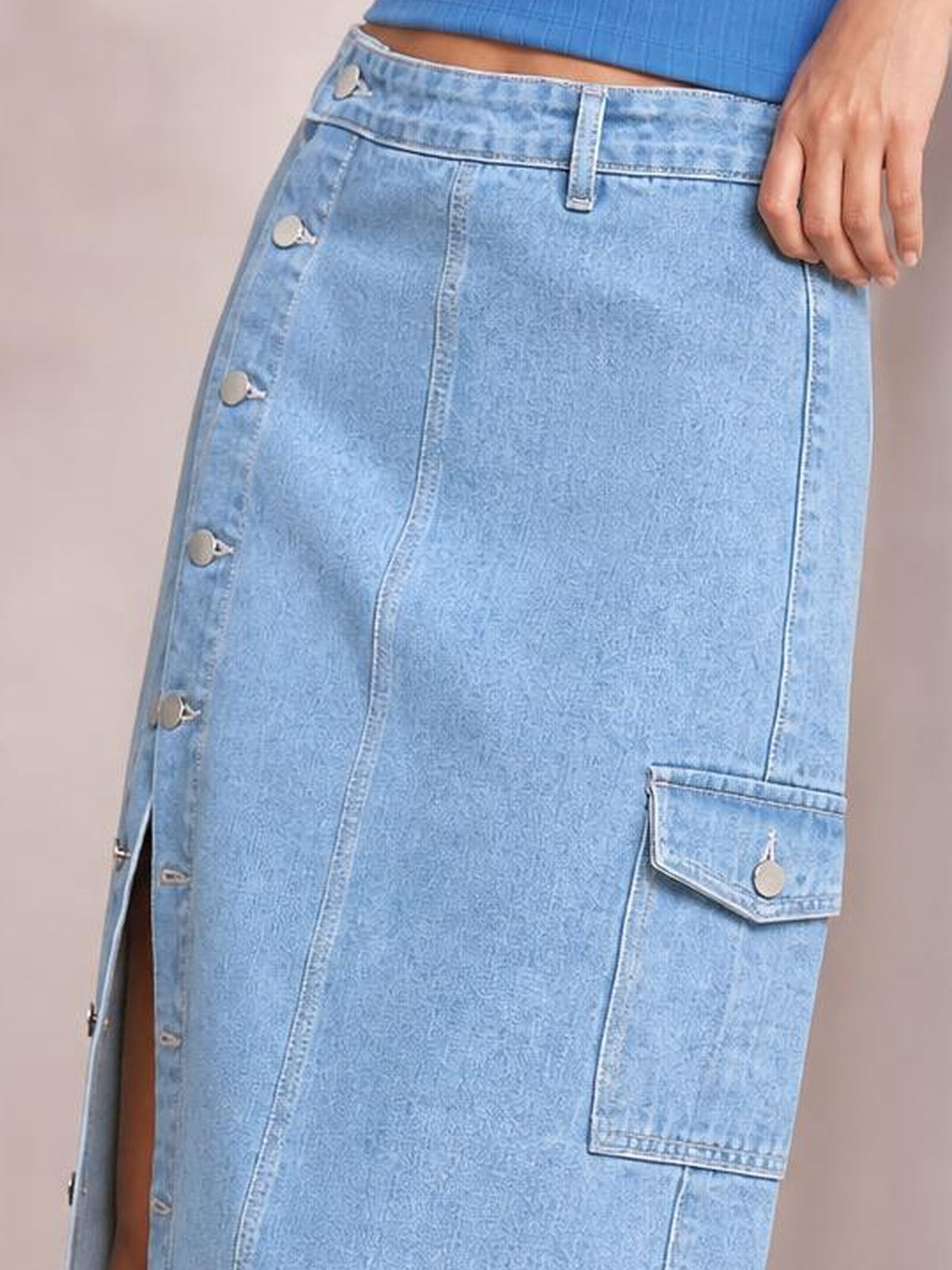 A cropped model stands with one hand at her hip, wearing a light-blue denim midi skirt with a straight silhouette, button-down front, side slit, and flap cargo pocket.