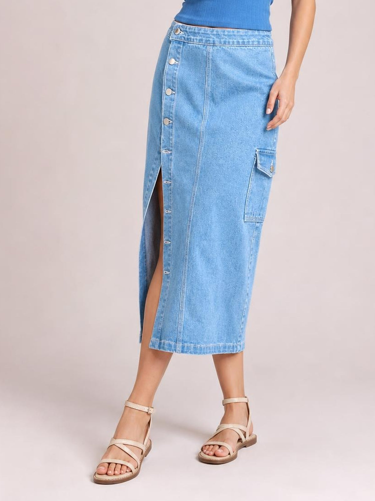 A model poses in a light blue button-down midi denim skirt with a straight silhouette, front slit, visible buttons, and a flap cargo pocket.
