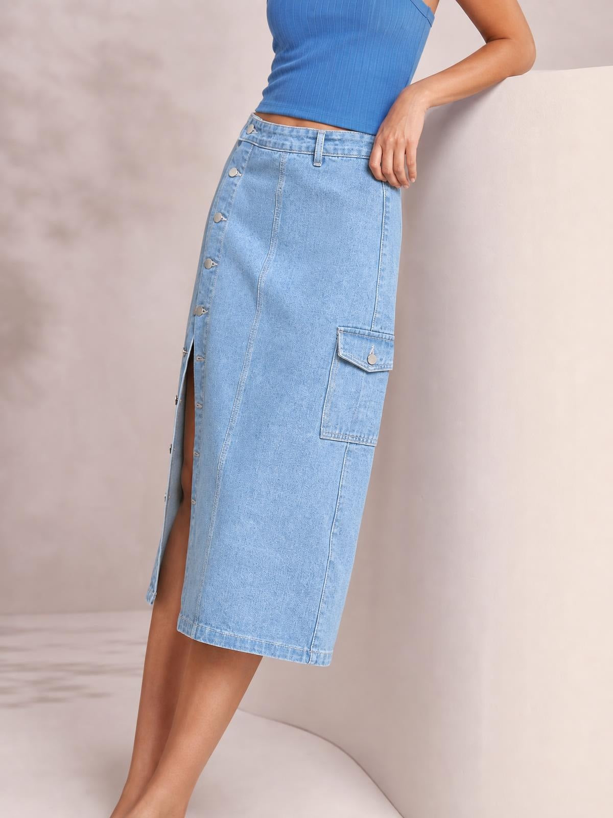 Model leans with one hand on a ledge, showcasing a light blue denim midi skirt with a straight silhouette, button-down front, side slit, and flap pocket.
