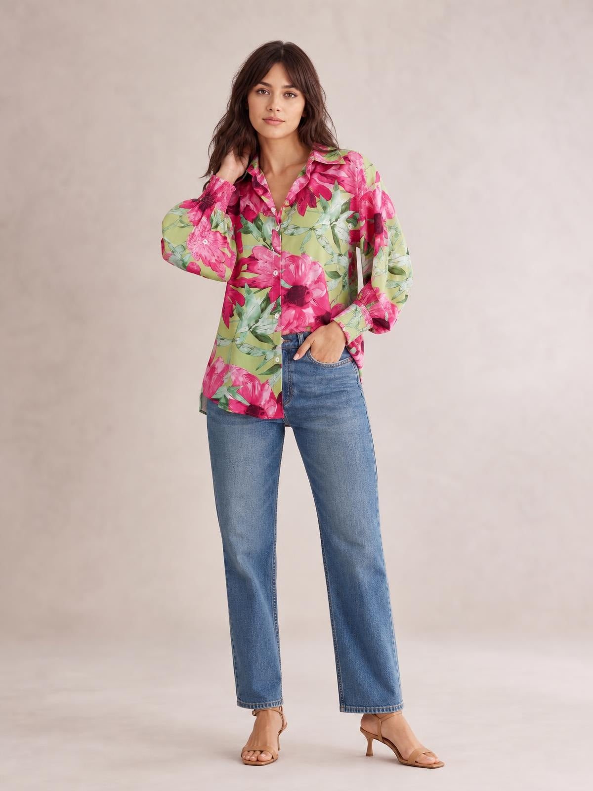 A model poses in a vibrant pink, green, and cream floral button-down long sleeve shirt with a collar, front buttons, relaxed silhouette, and one hand in her jeans pocket.