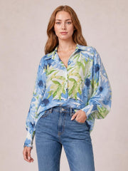 A model wears a blue and green floral button-down long sleeve shirt with a collared neckline, relaxed silhouette, and gathered cuffs, posing with one hand in her pocket.