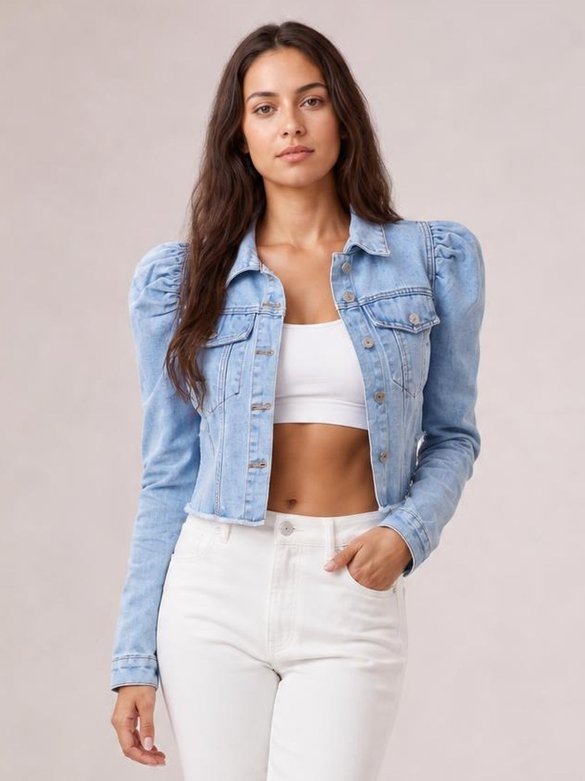 Model wearing a light blue button-down cropped denim jacket with puffed shoulders, long sleeves, chest pockets, and a raw hem, standing with one hand in her pocket.