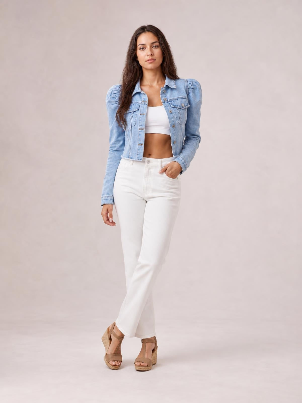 A model poses with one hand in her pocket, wearing a light blue button-down cropped denim jacket with puff sleeves, chest pockets, and a raw hem.