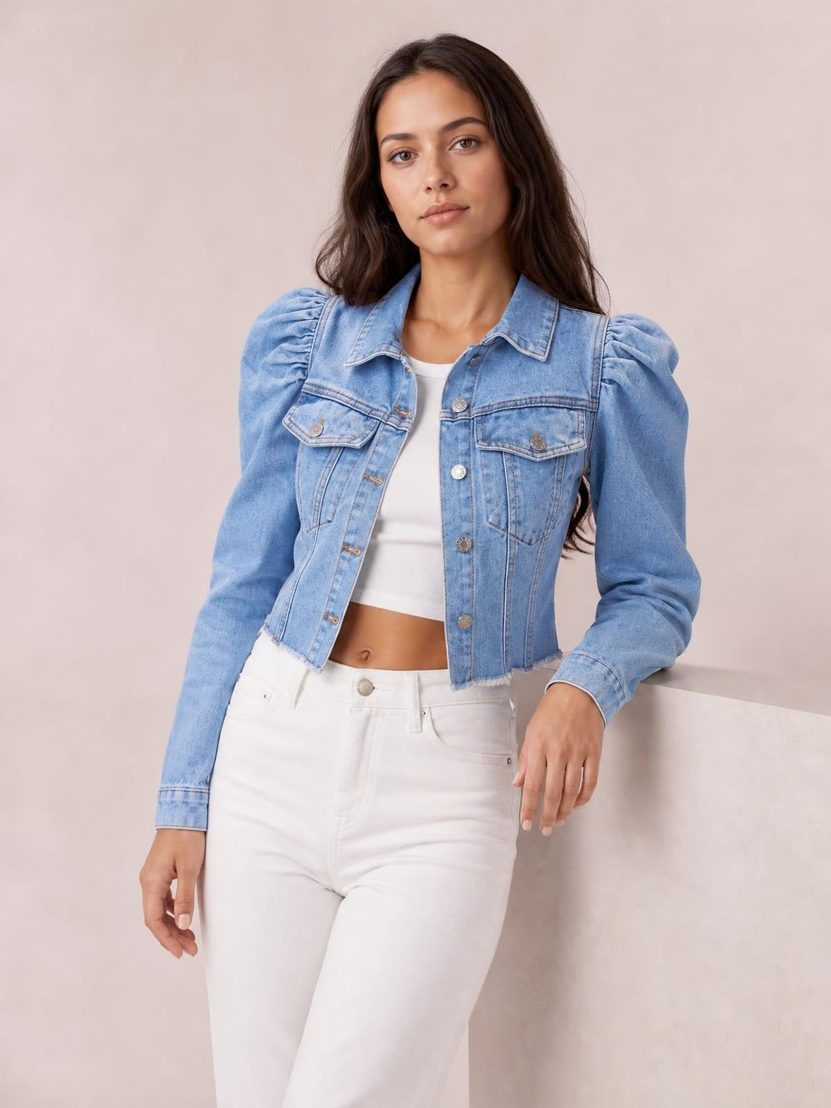 Model leans against a ledge wearing a light blue button-down cropped denim jacket with raw hem, puff sleeves, and front pockets.