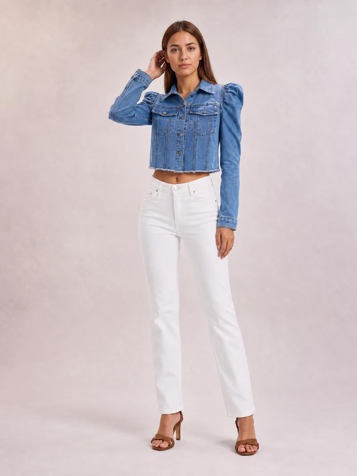 A model poses with one hand by her hair, wearing a blue button-down cropped denim jacket with puffed sleeves, chest pockets, structured seams, and a raw hem.