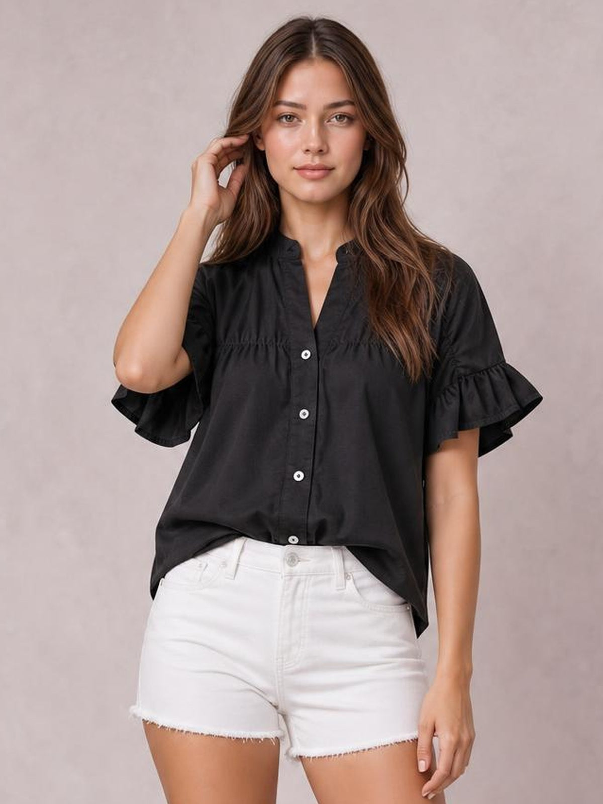 A model touches her hair while wearing a black button-down shirt with ruched detailing, a relaxed silhouette, and ruffled short sleeves.